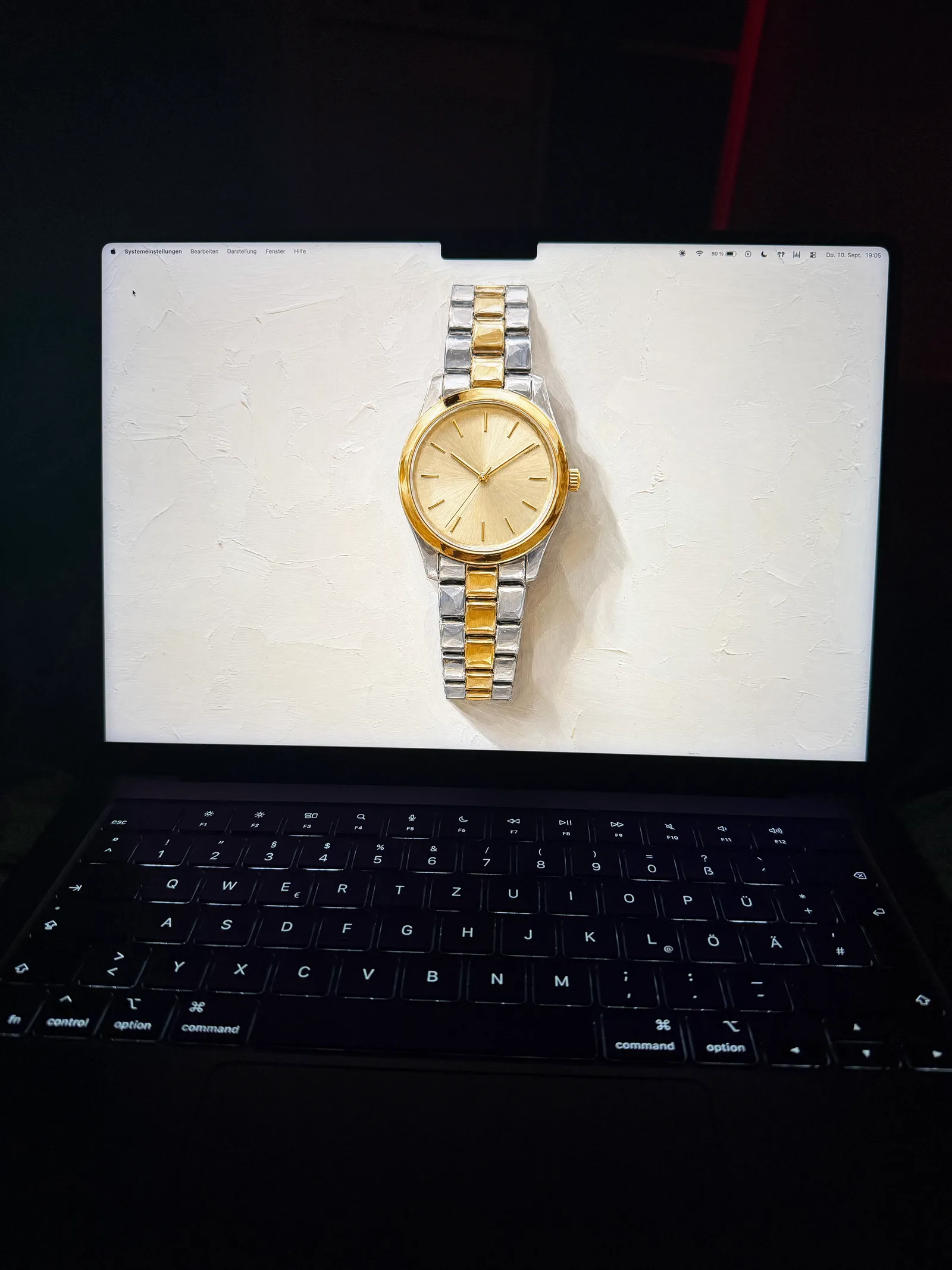 MacBook im abgedunkelten Raum, auf dem Bildschirm die goldene Armbanduhr auf hellem Impasto-Grund.